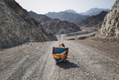 Woman holding map while sitting on road by mountains against sky