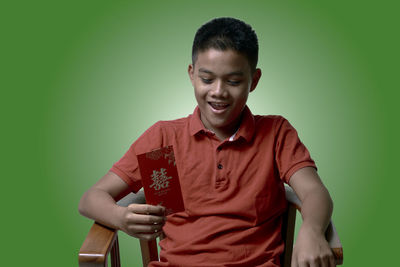 Smiling boy holding envelop while sitting on chair against green background
