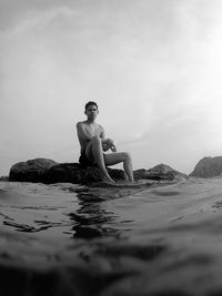 Shirtless man sitting on rock amidst sea against sky