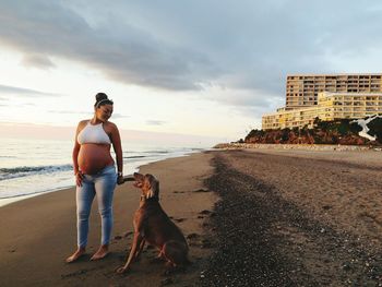 Full length of pregnant woman with dog on beach against sky