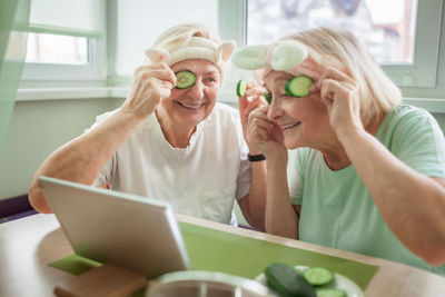 Two senior women apply facial cosmetic mask, have fun at home, make selfie and chat in social media