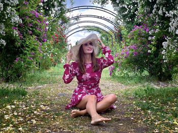 Woman in pink dress and sun hat sitting down on the ground in a garden of roses