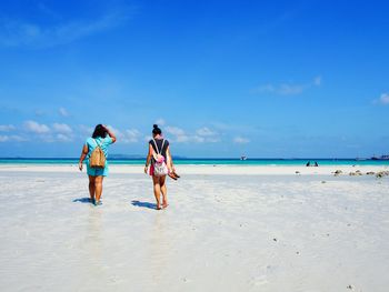 Rear view full length of women walking at sandy beach against blue sky