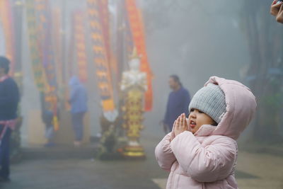 Cute girl wearing warm clothing praying while standing during foggy weather