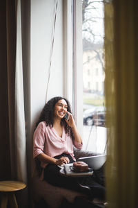 Young woman using mobile phone while sitting on window