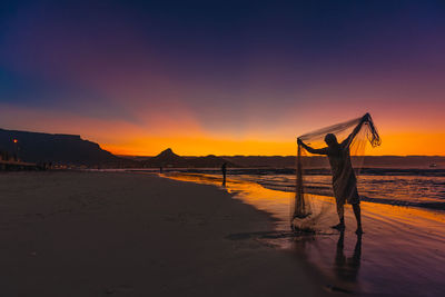 Silhouette man holding up a fishing net at beach during sunset and table mountain in the background 