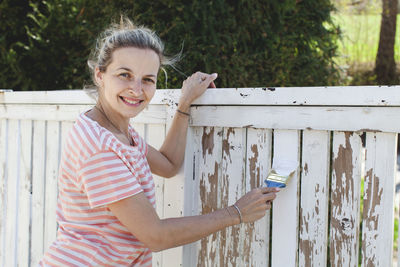 Portrait of smiling woman painting fence at yard
