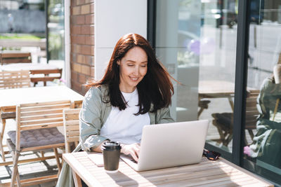 Adult smiling brunette business woman forty years in stylish shirt working on laptop in cafe