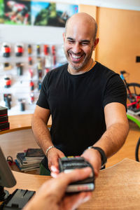 Delighted adult man in black t-shirt smiling and giving payment terminal to crop client while working in bike shop