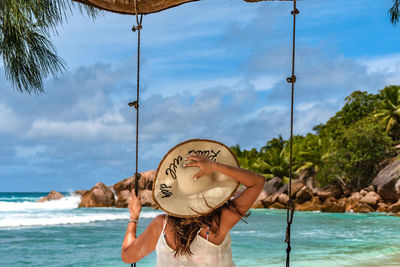 Rear view of young woman wearing beach clothes, sitting on swing on paradise beach