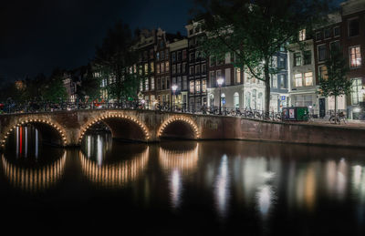 Illuminated bridge over river by buildings in city at night