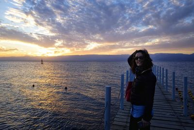 Portrait of smiling woman standing on pier over sea against cloudy sky during sunset