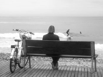 Rear view of woman sitting on bench while looking at man surfing on sea