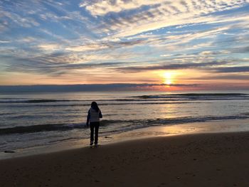 Rear view of woman standing on beach by sea against sky during sunset