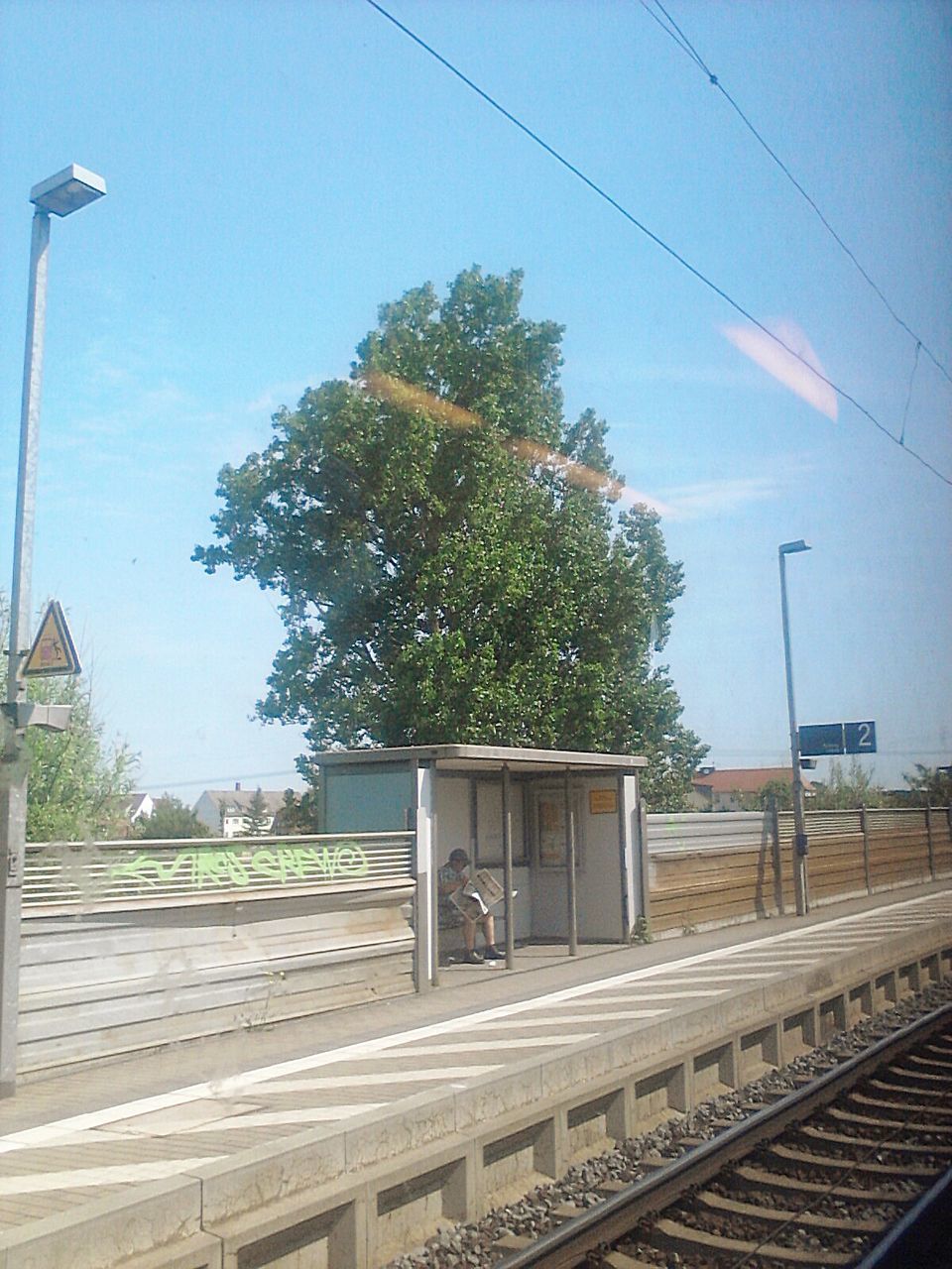 railroad track, rail transportation, power line, tree, clear sky, transportation, built structure, sky, architecture, public transportation, cable, building exterior, street light, electricity pylon, railroad station, electricity, connection, railroad station platform, power supply, blue