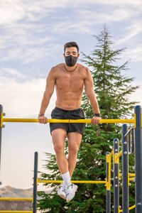 Full length of shirtless man sitting on railing against sky