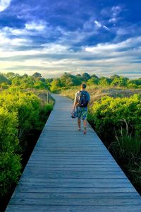 Rear view of man walking on boardwalk against sky