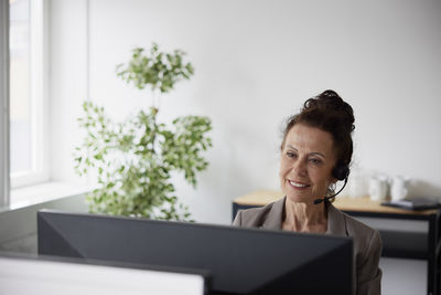 Smiling mature businesswoman using headset in office in front of computer screen
