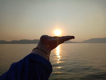 Person hand by sea against sky during sunset