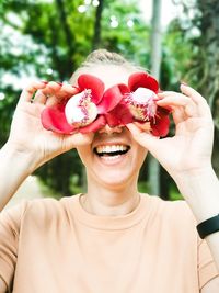 Smiling young woman holding flowers over eyes against trees