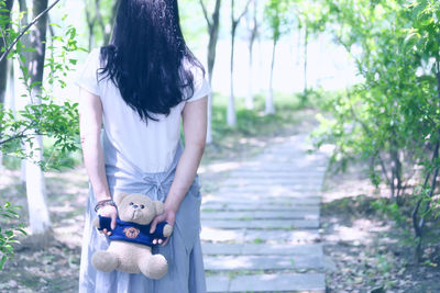 Rear view of woman holding teddy bear behind back while walking on walkway in park
