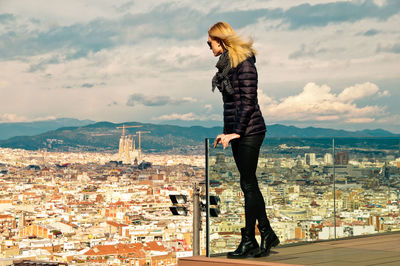 Young woman wearing warm clothing while standing on balcony against cityscape