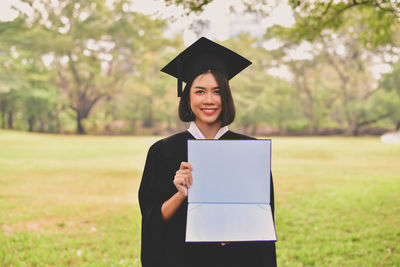 Portrait of young woman in graduation gown holding file while standing at park