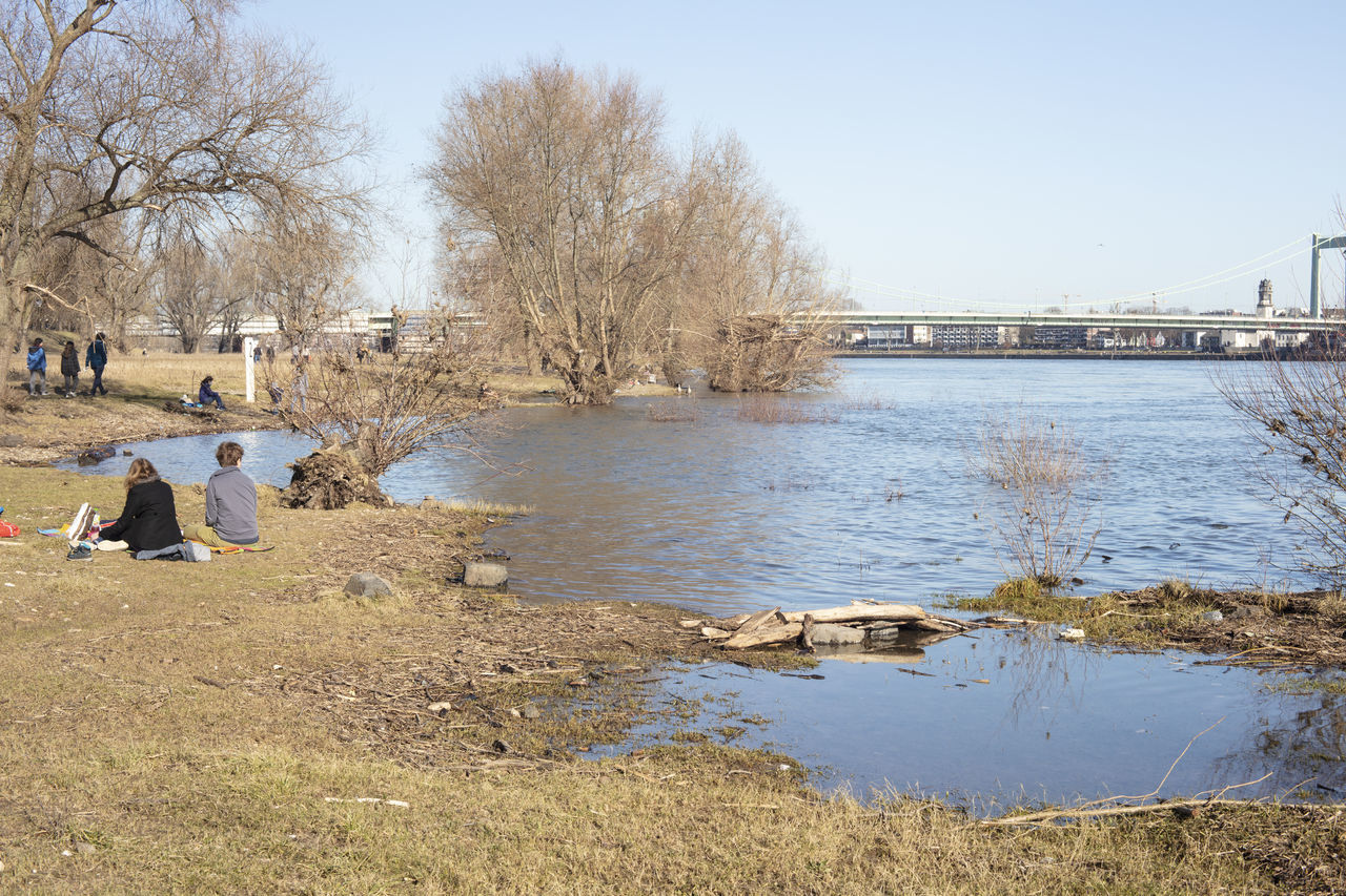 PEOPLE SITTING ON RIVERBANK BY RIVER AGAINST SKY