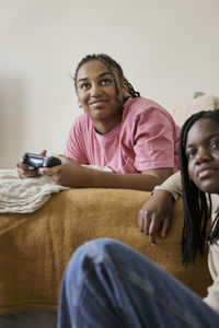 Teenage girl playing video game by female friend in bedroom during weekend