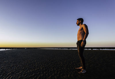 Young shirtless man looking at sea against sky during sunset