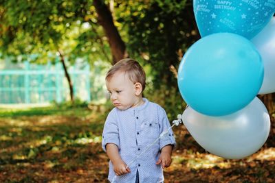 Cute baby boy with balloons standing against trees