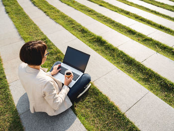 Business woman sits in park with laptop and take away cardboard cup of coffee. urban lifestyle.