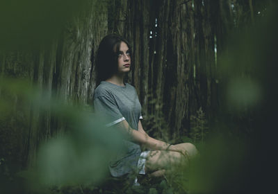Side view of young woman sitting against tree trunk in forest