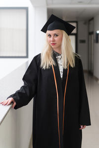 Portrait of young woman wearing graduation gown standing against wall