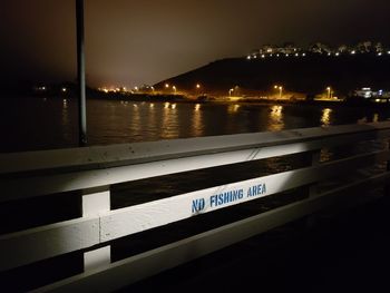 Illuminated sign by river against sky at night
