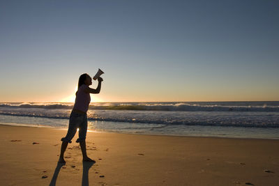 Side view of mid adult woman standing at beach against clear sky during sunset