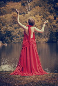 Full length rear view of young woman in red evening gown standing on grass at lakeshore