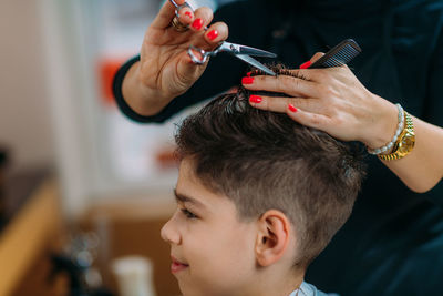 Boy getting a new haircut. female hairstylist cutting his black hair with scissors in hair salon