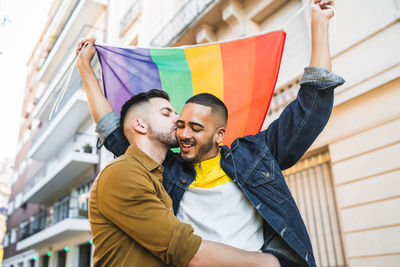 Gay couple holding rainbow flag while standing in city