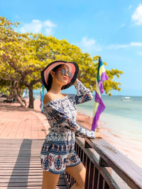 Young woman wearing sunglasses while standing by railing at beach against sky