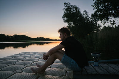 Side view full length of young man sitting by lake during sunset