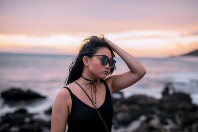 Young woman wearing sunglasses while standing at beach against sky during sunset