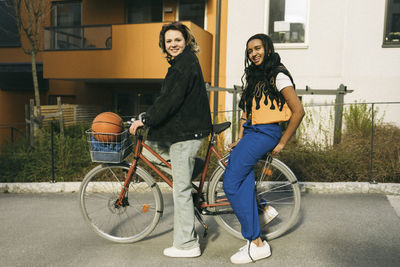 Portrait of smiling female friends sitting on bicycle in front of building at footpath