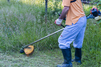 A mature man in protective clothing, and gloves  and mowing tall grass and the weeds with a trimmer