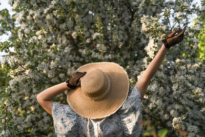 Woman in retro clothes with straw hat standing back in spring blossom apple tree garden.