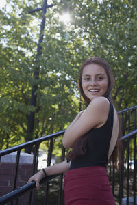 Portrait of smiling woman standing by railing against trees