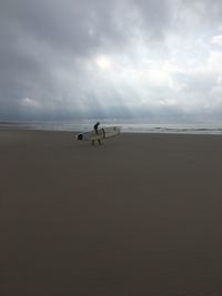 Side view of man with surfboard walking at beach against cloudy sky