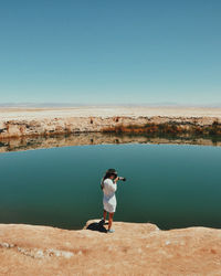 Full length of woman photographing calm lake against clear blue sky