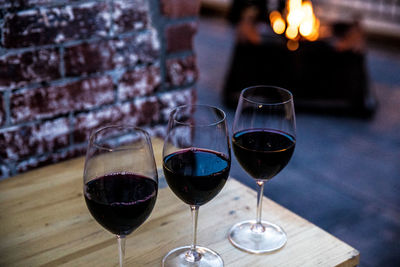 Three red wine glasses sitting on table with a brick wall and a glowing fire in the background