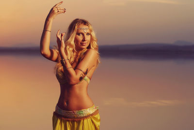 Portrait of beautiful young woman standing at beach against sky during sunset